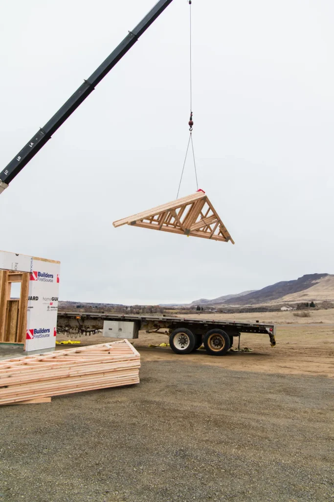 A crane lifting a single wooden triangular truss from a flatbed trailer at an Anasazi Builders job site.