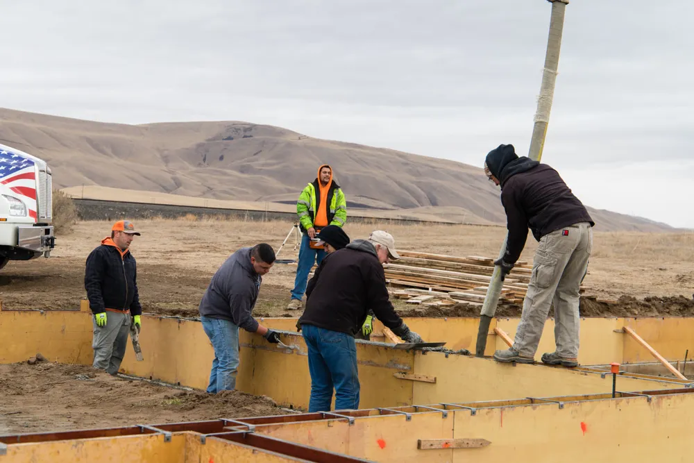 A close-up view of several construction workers meticulously leveling and smoothing freshly poured concrete into wooden foundation forms. One worker guides the discharge hose from a concrete pump, while others use hand tools for finishing, set against a backdrop of rolling hills under a cloudy sky, completed by Anasazi Builders in Tri-Cities.