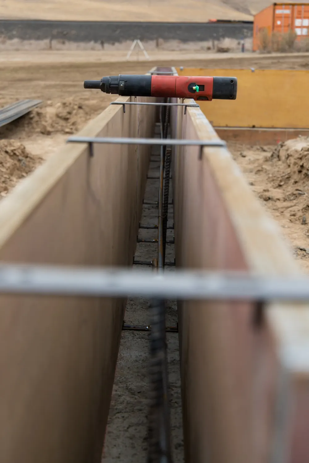 A close-up, ground-level view of a narrow concrete foundation trench being prepared with wooden forms and metal rebar. A red power tool sits on top of the formwork, and the background shows a dirt construction site with a distant orange shipping container, completed by Anasazi Builders in Tri-Cities.