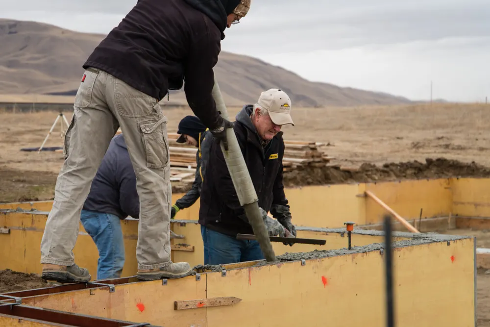 Two construction workers focusing on a concrete pour into yellow wooden foundation forms. One worker, standing on the edge of the forms, guides the pump hose while the other, wearing a tan cap, uses a hand tool to smooth the wet concrete, completed by Anasazi Builders in Tri-Cities.