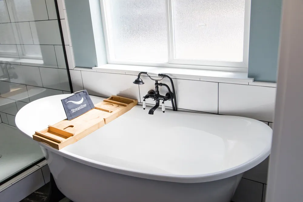 Luxury bathroom design by Anasazi Builders in Benton County, WA, featuring a white freestanding Streamline soaking tub, vintage-style black faucet fixtures, and a bamboo bathtub caddy tray against a white subway tile backdrop.