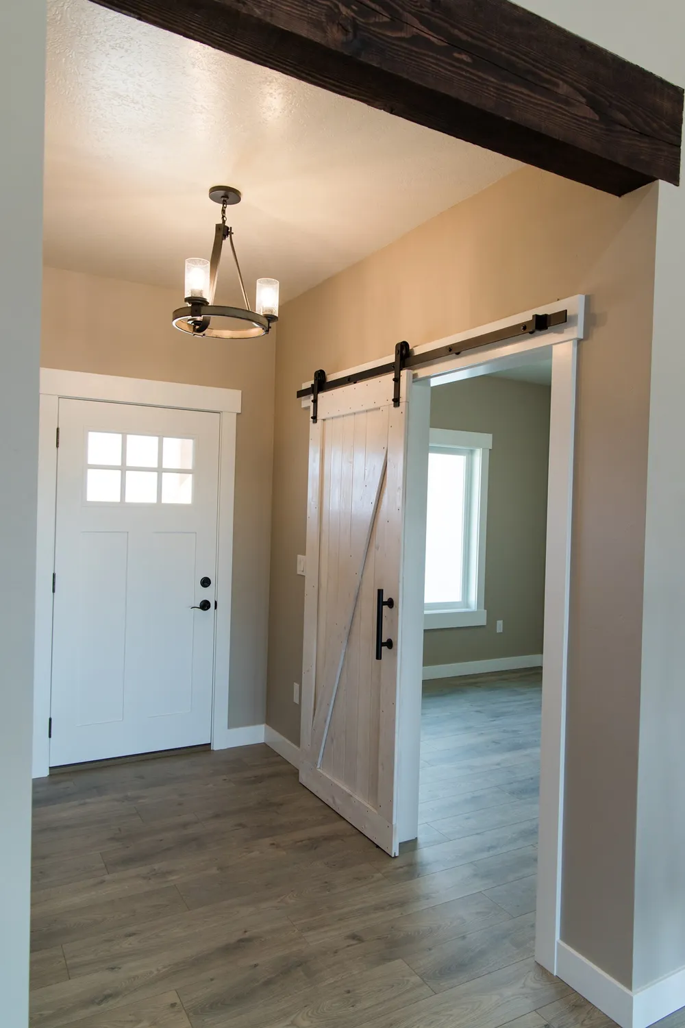 Interior entryway of a modern home featuring a white craftsman-style front door with a window and a white sliding barn door with black hardware, leading to a room with light wood flooring and a large window, installed by Anasazi Builders in Tri-Cities.
