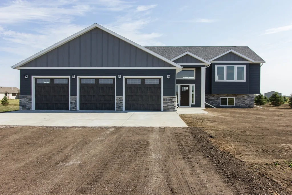 Exterior view of a new custom home built by Anasazi Builders in Benton County, WA. The home features dark grey vertical and horizontal siding, a stone veneer foundation, and a three-car garage with wood-grain doors, set against a rural landscape.