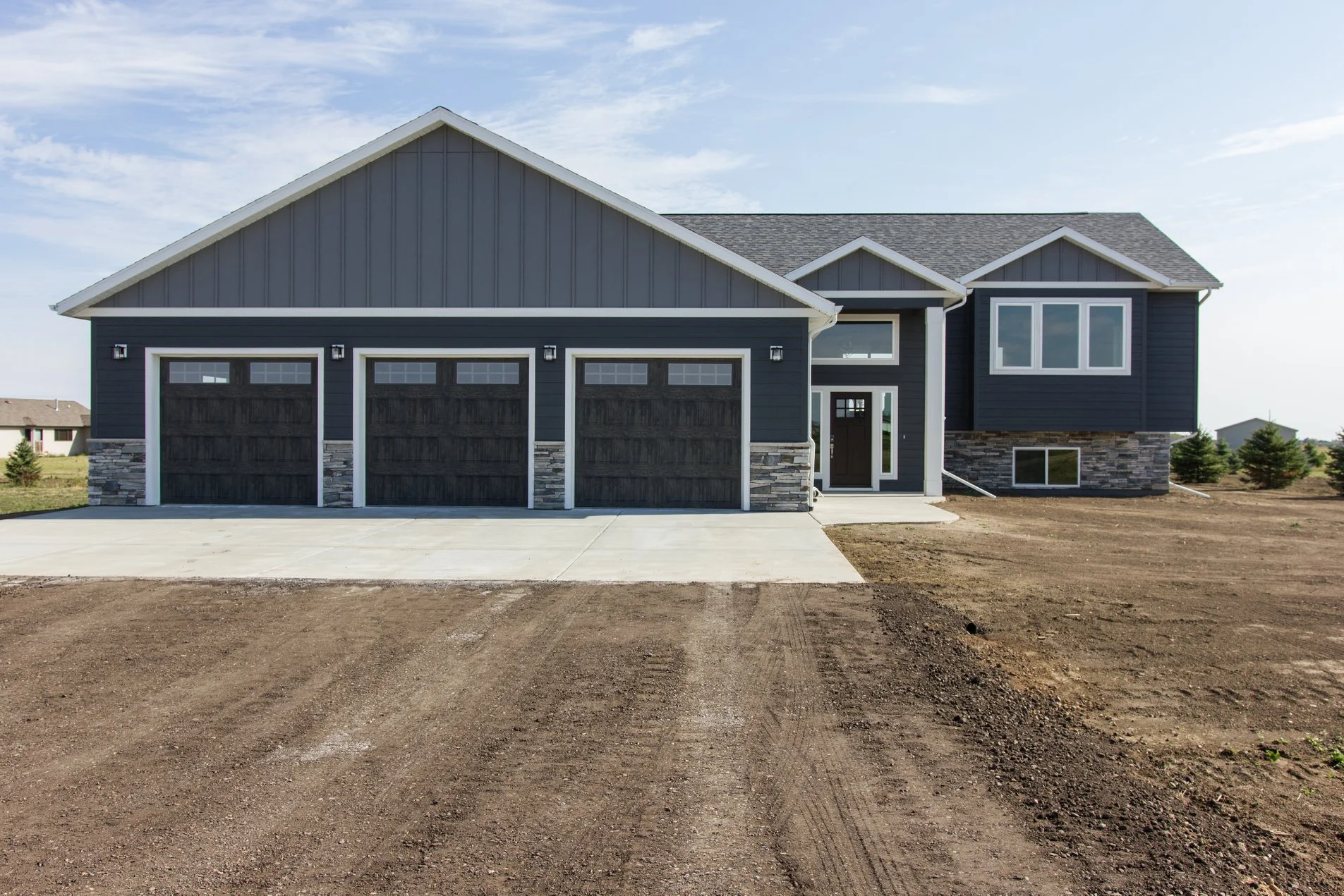 Exterior view of a new custom home built by Anasazi Builders in Benton County, WA. The home features dark grey vertical and horizontal siding, a stone veneer foundation, and a three-car garage with wood-grain doors, set against a rural landscape.