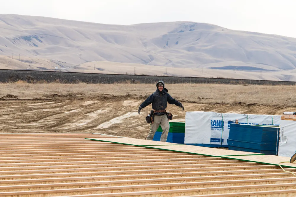 A professional builder from Anasazi Builders installing subflooring on a new custom home in Tri-Cities, featuring wooden floor joists and a scenic hill background.