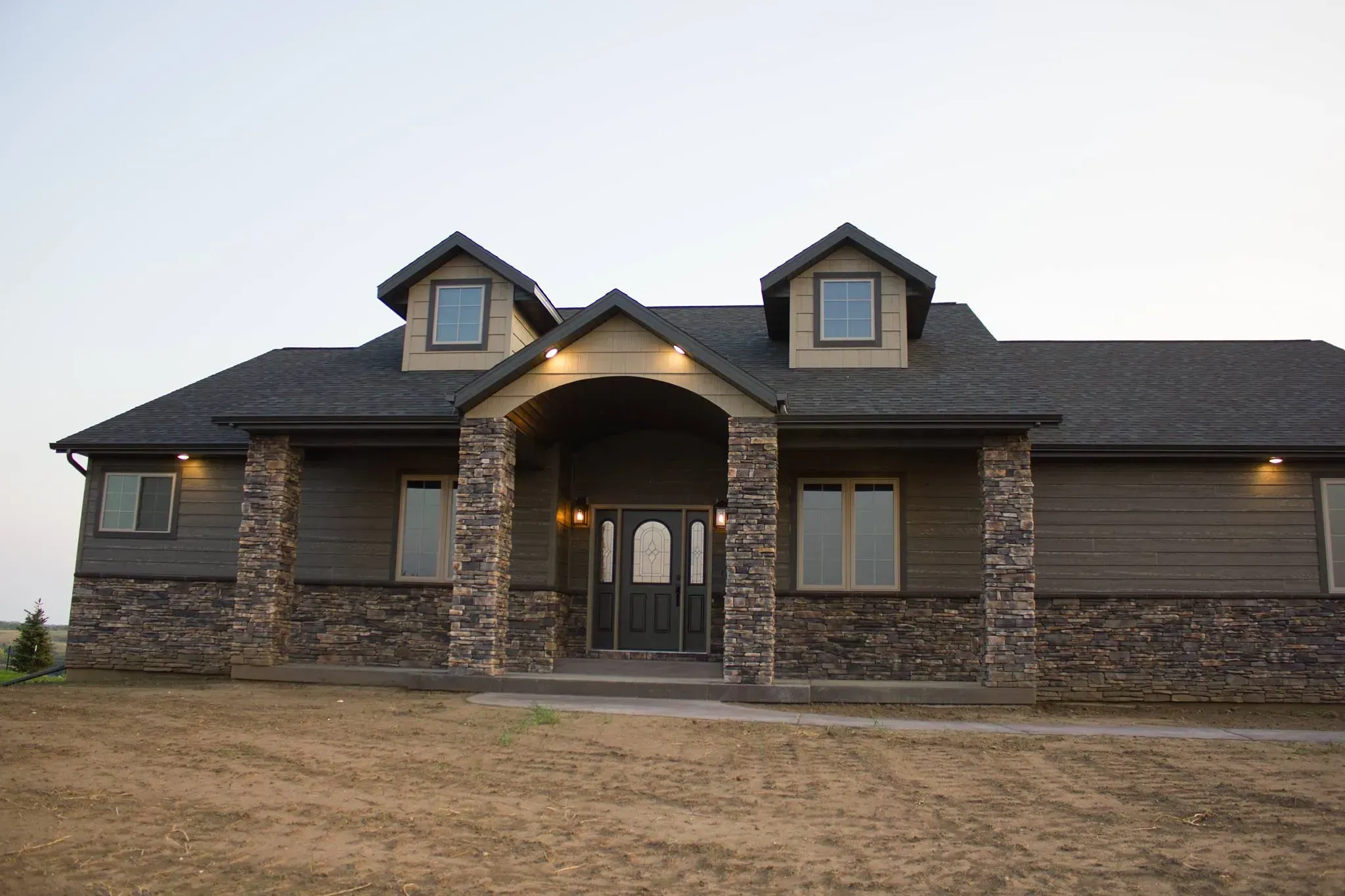Exterior front view of a custom ranch-style home with grey siding, extensive stone masonry, a vaulted entryway, and two dormer windows, by Anasazi Builders.