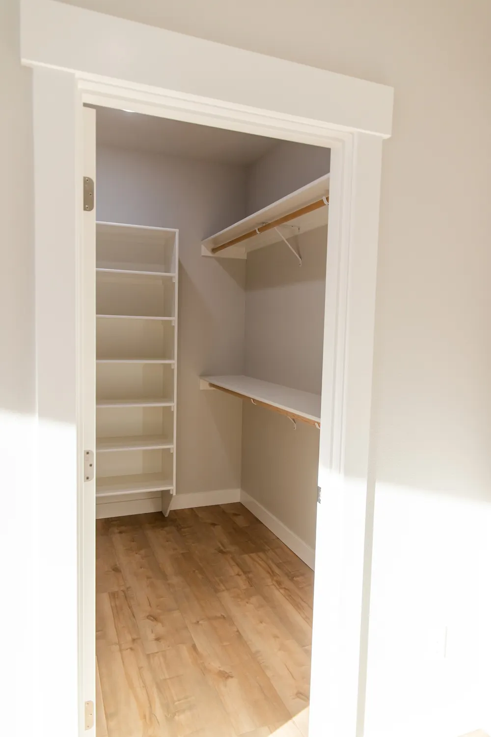 A view through a white craftsman-style door frame into a walk-in closet featuring light wood flooring, a set of six white built-in shelves on the left, and two long white hanging rods with upper shelving, completed by Anasazi Builders in Tri-Cities.