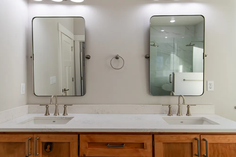 A close-up view of a master bathroom double vanity featuring a natural wood cabinet, a white quartz countertop with two undermount sinks, and two large rectangular pivot mirrors, completed by Anasazi Builders in Tri-Cities.