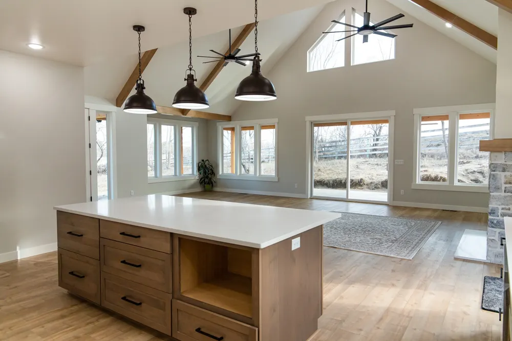 A finished open-concept great room featuring a modern kitchen island with natural wood cabinets, three industrial black pendant lights, and a vaulted ceiling with exposed wood beams and dual ceiling fans, completed by Anasazi Builders in Tri-Cities.