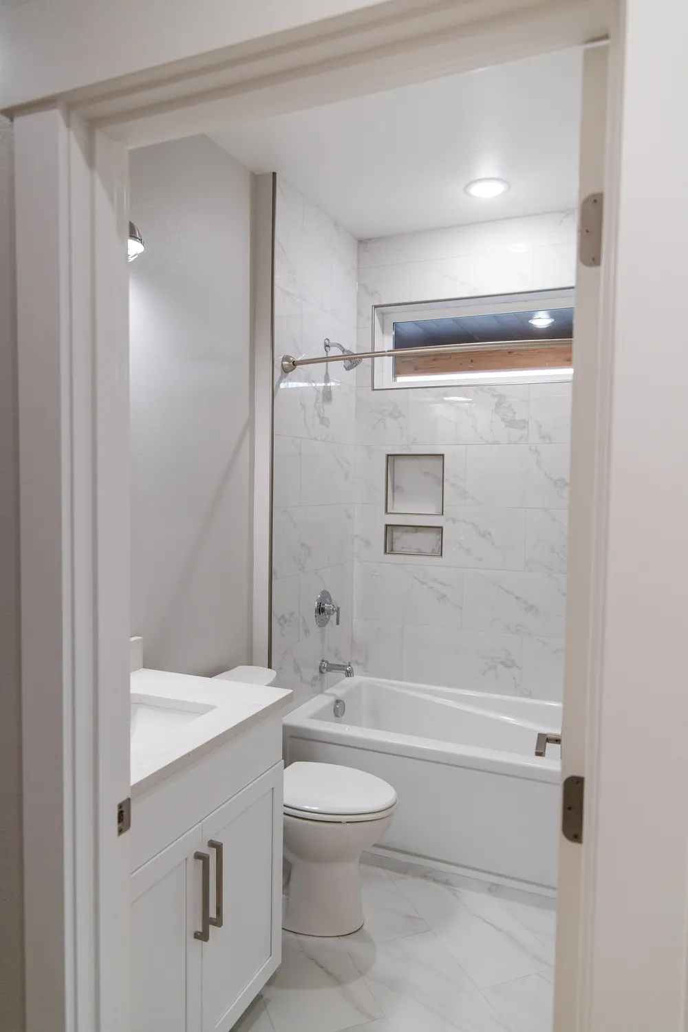 A view into a bright guest bathroom featuring a white vanity with chrome hardware, a white toilet, and an alcove bathtub with marble-patterned tile walls and two recessed storage niches, completed by Anasazi Builders in Tri-Cities.