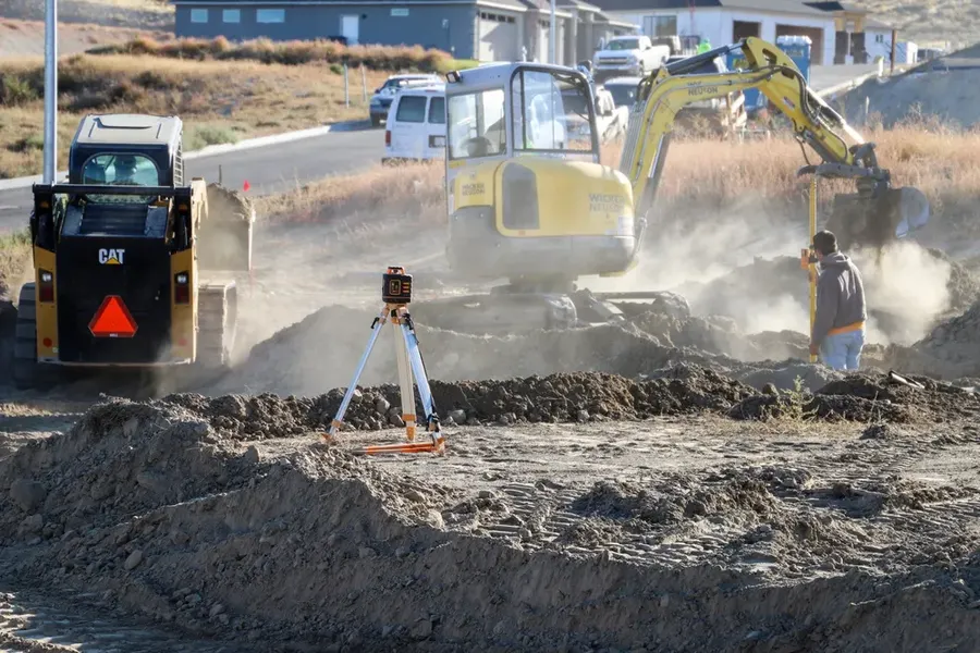 A yellow CAT 259D skid steer loader and a yellow Wacker Neuson excavator are engaged in earthmoving activities at a dusty residential construction site. A laser level is set up on a tripod in the foreground, and a worker is visible near the excavator as dust rises from the ground, with newly built homes in the background under a clear sky, completed by Anasazi Builders in Tri-Cities.