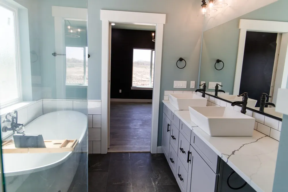 A finished master bathroom featuring a freestanding white bathtub with a wooden tray, a double vanity with grey cabinets and vessel sinks, and teal walls, completed by Anasazi Builders in Tri-Cities.