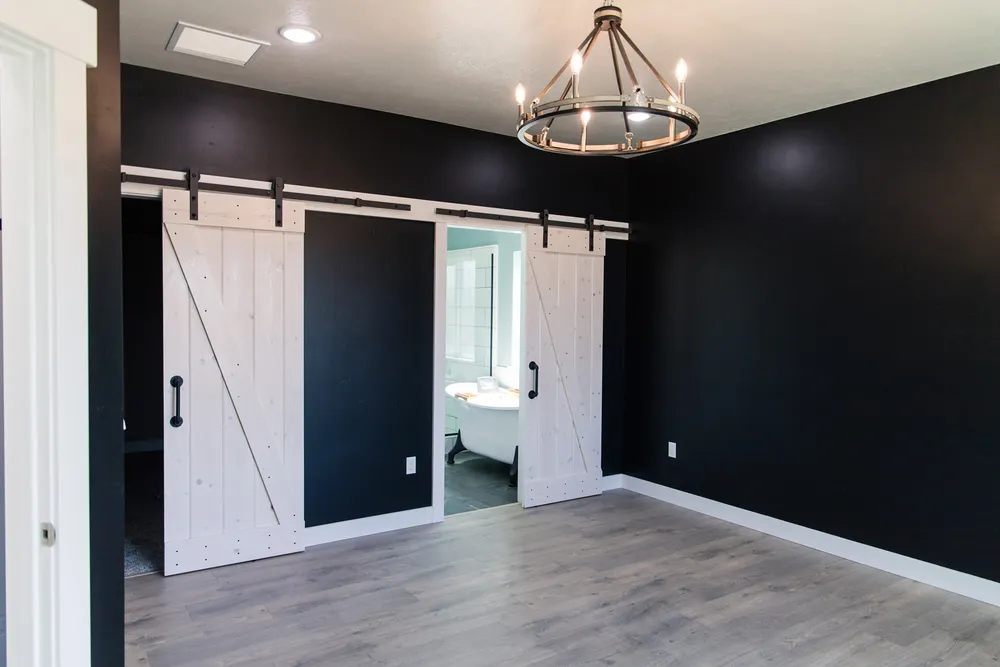 A finished bedroom with black walls and white baseboards featuring two white sliding barn doors with black hardware, providing access to a master bathroom with a freestanding tub, and a modern circular chandelier, completed by Anasazi Builders in Tri-Cities.