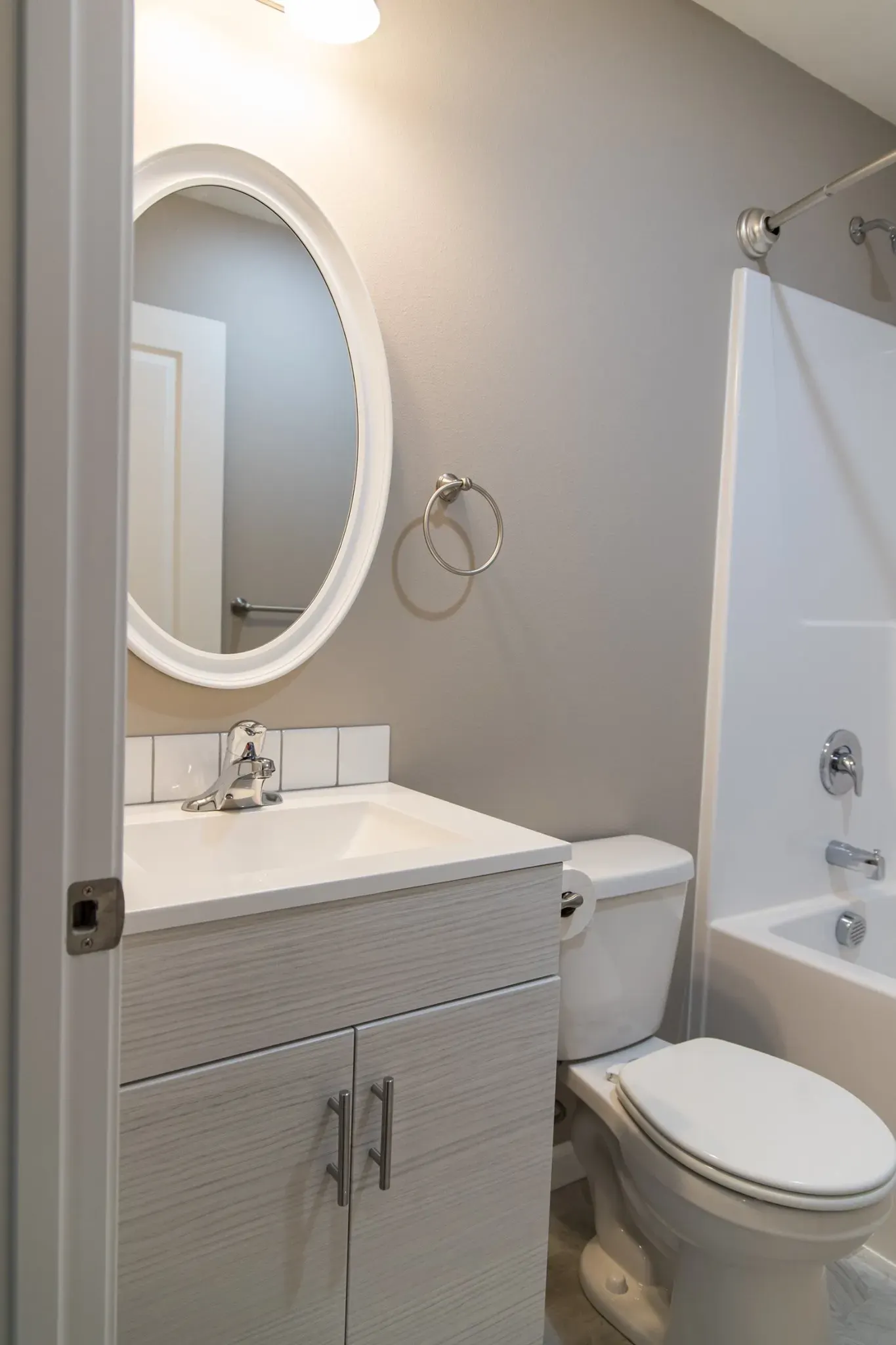 Modern bathroom featuring a light wood-textured vanity with a white square sink, an oval mirror, a white toilet, and a bathtub with brushed nickel fixtures.