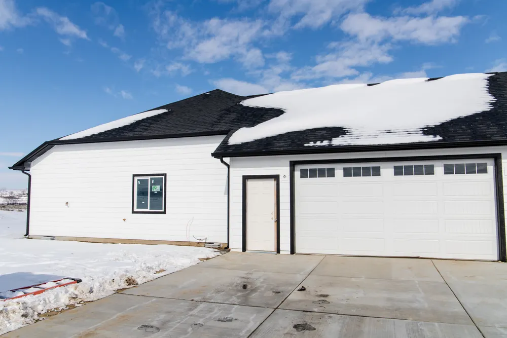 Side view of a white modern farmhouse garage with black trim, a white side door, and a large sectional garage door on a concrete driveway with snow by Anasazi Builders in Tri-Cities.