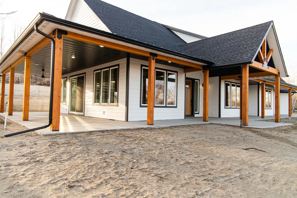 A perspective view of a modern farmhouse exterior featuring a large covered patio with natural wood pillars, white horizontal siding, and black trim, leading to the gabled front entrance with exposed wood trusses, completed by Anasazi Builders in Tri-Cities.