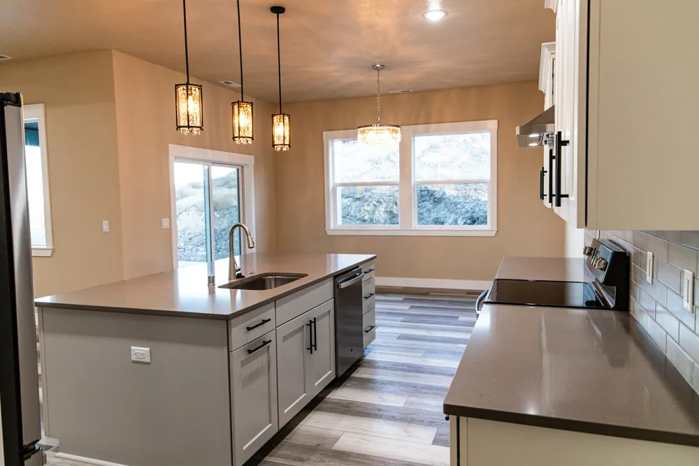 Modern kitchen with a grey center island, stainless steel sink, and three elegant gold pendant lights, looking toward an open dining area, by Anasazi Builders in Tri-Cities.