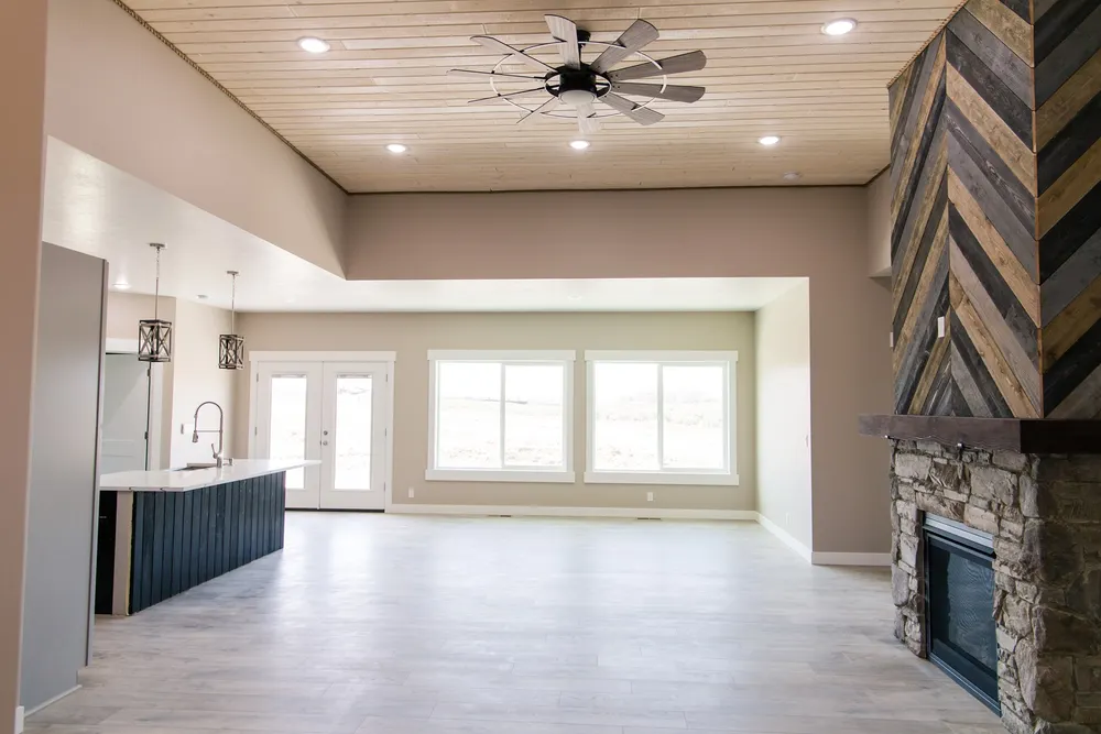 A wide open-concept living area featuring a stone fireplace with a wood chevron mantel, a kitchen island with a dark base, and a wood plank ceiling with a large fan, built by Anasazi Builders in Tri-Cities.