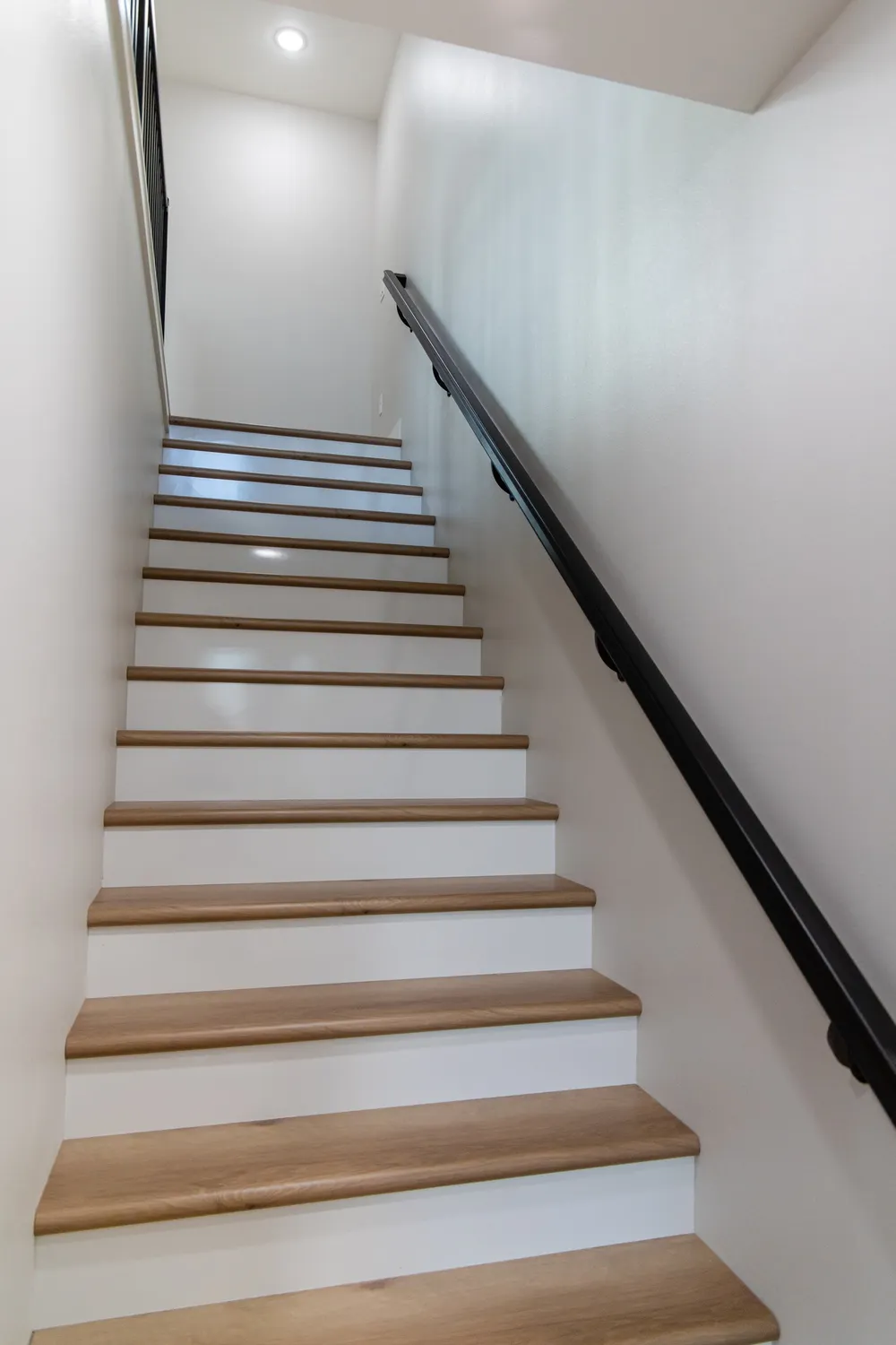 A straight indoor staircase featuring white risers and light wood treads, accompanied by a sleek black handrail mounted on a light grey wall, leading to a second-floor landing with recessed lighting, completed by Anasazi Builders in Tri-Cities.