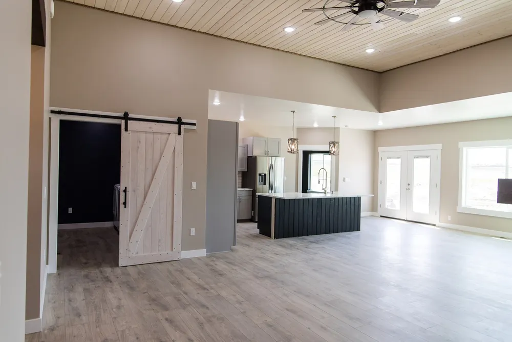 A wide-angle view of a modern open-concept living area featuring light wood-look flooring, a kitchen with a dark-base island and white countertops, and a white sliding barn door leading to a laundry room with navy blue walls, built by Anasazi Builders in Tri-Cities.