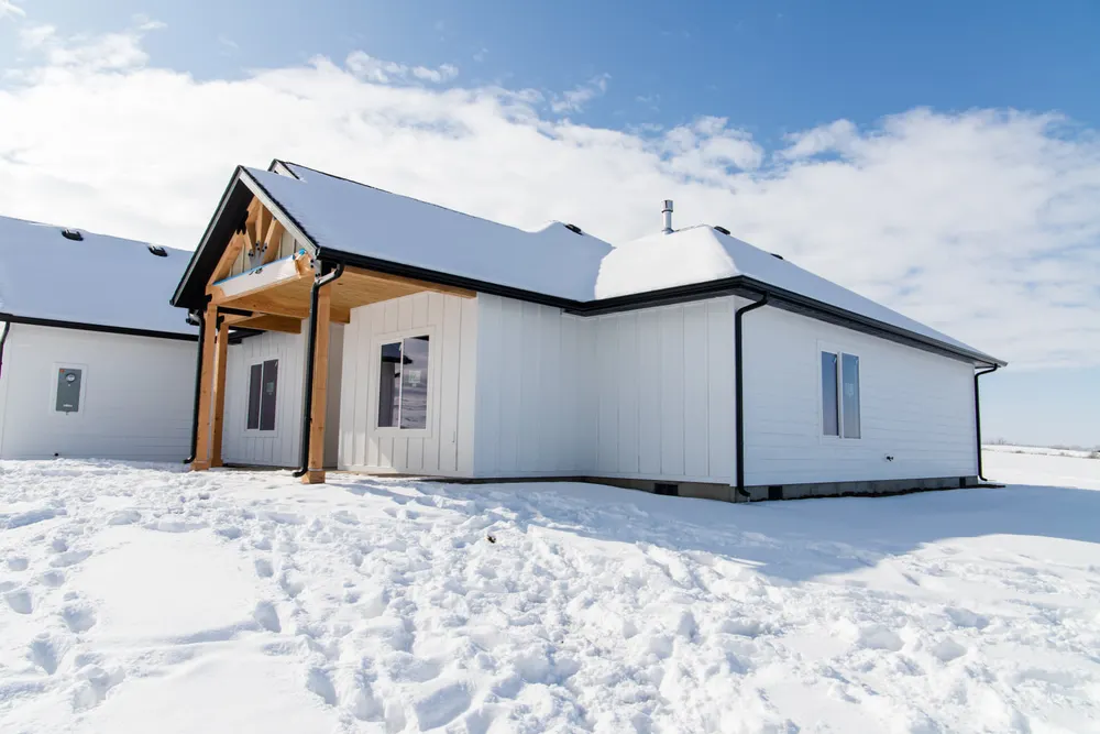 Exterior of a white modern farmhouse with black gutters and wood porch pillars, partially covered in snow on a sunny day in Benton County, built by Anasazi Builders.