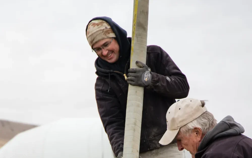 A construction worker in a grey hoodie and a dark beanie stands next to yellow wooden foundation forms. He is holding a tool and looking down toward the foundation, with a white concrete mixer truck and dry, rolling hills in the background under an overcast sky, completed by Anasazi Builders in Tri-Cities.