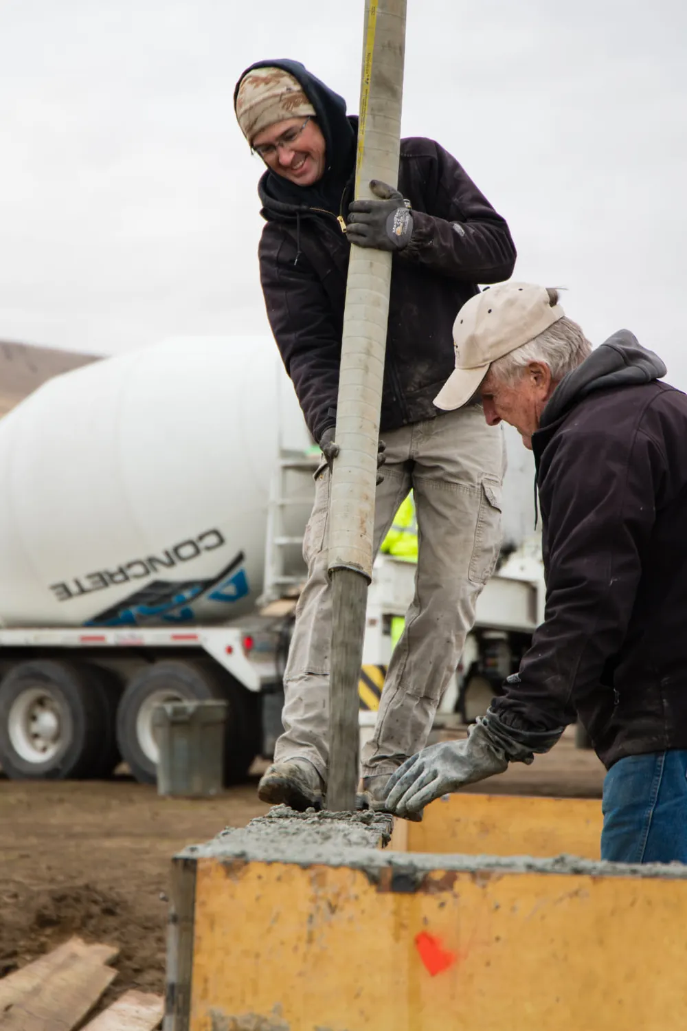 A construction worker in a grey hoodie and a dark beanie stands next to yellow wooden foundation forms. He is holding a tool and looking down toward the foundation, with a white concrete mixer truck and dry, rolling hills in the background under an overcast sky, completed by Anasazi Builders in Tri-Cities.