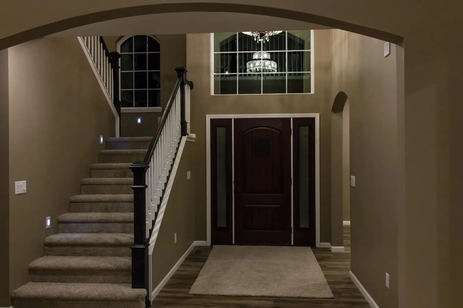 Grand foyer interior featuring a dark wood front door with sidelights, a carpeted staircase with dark railings, and a view of a crystal chandelier through an upper window.
