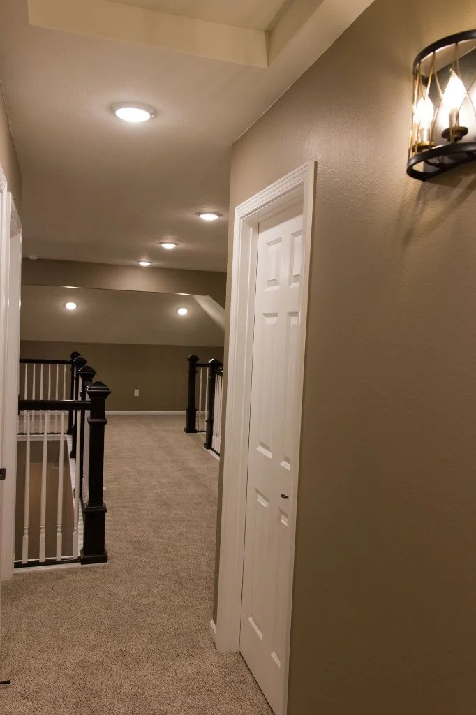Second-floor hallway of a custom home featuring beige carpeting, tan walls, a white paneled door, and a decorative black and gold wall sconce.