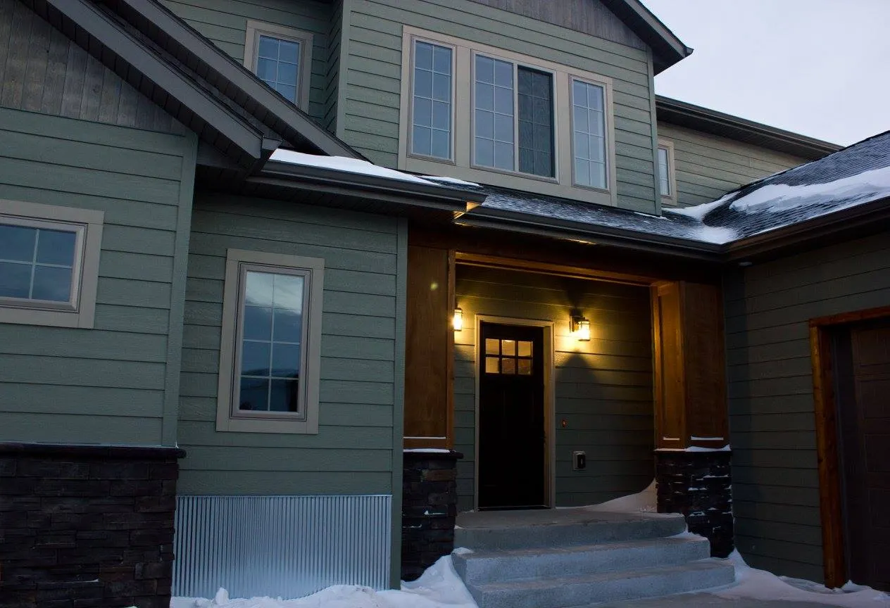 Exterior view of a modern two-story house featuring olive green lap siding, dark stone accents, and wood-wrapped columns. The entrance has a dark front door with window panes, illuminated by two warm outdoor wall lights, with concrete steps covered in light snow