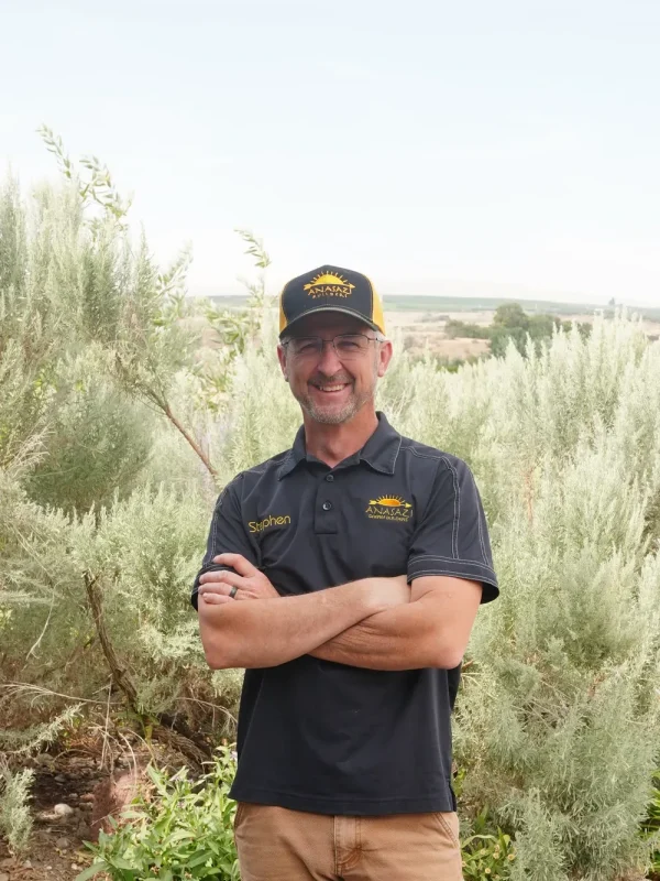 A smiling portrait of Stephen Wourland, owner of Anasazi Builders, wearing a branded black polo shirt and cap with crossed arms, standing outdoors.